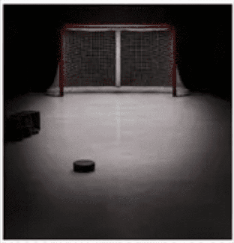 A hockey puck in front of an empty hockey goal on ice.